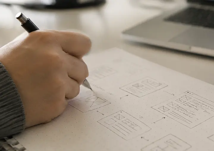 Mão rascunhando telas de um site num caderno, ao lado de um notebook e de uma caneca